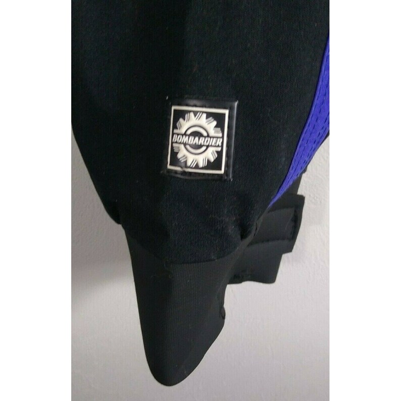 May include: Close-up of a black garment with a blue mesh accent. A square patch with the word "BOMBARDIER" and a gear design is visible. The garment has a ribbed cuff and a smooth, matte finish.