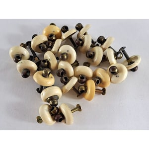 Könnte beinhalten: Eine Sammlung von 18 cremefarbenen Keramik-Schubladengriffen mit Messing-Schrauben. Die Griffe sind rund und haben eine erhabene Mitte.