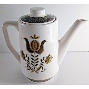 Può includere: Una caffettiera in ceramica bianca con un motivo floreale marrone e oro. La caffettiera ha un coperchio con un motivo a pois marroni.