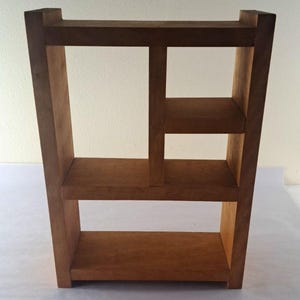 May include: A wooden bookshelf with three shelves and a vertical support in the center. The bookshelf is made of light brown wood and has a simple, geometric design. The shelves are rectangular and the overall structure is sturdy.