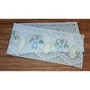 Puede incluir: Camino de mesa de encaje blanco con diseños florales y de tetera. El camino de mesa presenta un patrón repetido de flores rosas y verdes en jarrones azules y teteras amarillas. Los bordes son festoneados y el encaje tiene un tejido delicado y abierto.