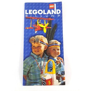 May include: A colorful brochure for Legoland Billund, featuring two children wearing headbands and a red and yellow airplane. The brochure has a blue background with white circles.