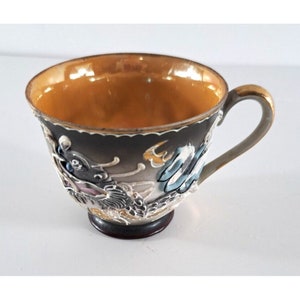 May include: A small, ceramic teacup with a gold rim and handle. The cup is decorated with a detailed, embossed design of a dragon in shades of gray, blue, and pink.