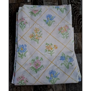 May include: White bed sheet with a repeating pattern of embroidered flowers in pink, blue, and yellow. The flowers are arranged in a grid pattern with gold dotted lines. The sheet is folded.