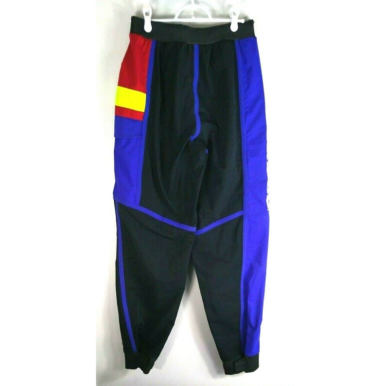 May include: Black, blue, red, and yellow athletic pants hanging on a white hanger. The pants have a black base with blue side panels and red and yellow accents. The pants have an elastic waistband and are made of a lightweight material.