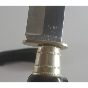 May include: Close-up of a silver knife handle with the words "Flint Vanadium USA" engraved on the side. The handle has a decorative silver band with a black handle.