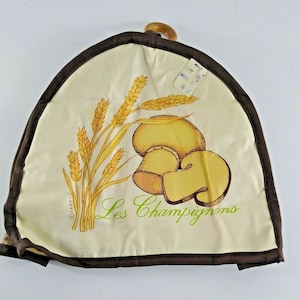 以下が含まれることがあります： ベージュの生地のパンカバーで、茶色の縁取りと木製の取っ手があります。カバーには、小麦の穂とキノコのデザインが施され、緑色の文字で「Les Champignons」と書いてあります。