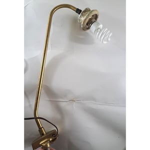 May include: A vintage brass desk lamp with a curved arm and a white, spiral-shaped light bulb. The lamp has a small base and a cord for plugging into an outlet. The lamp is designed to provide focused light for reading or working.