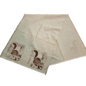 May include: A cream-colored scarf with a repeating pattern of brown cat illustrations. The scarf has a fringed edge and a simple, minimalist design. The cat illustrations are in a blocky, stylized design.