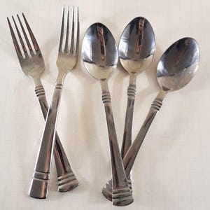 May include: A collection of stainless steel silverware, including two forks and three spoons. The forks have four tines, and all pieces have a decorative band near the handle. The silverware is arranged on a white surface.