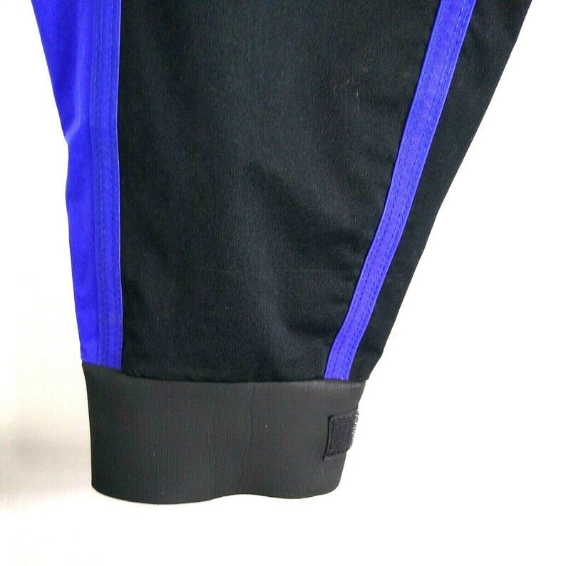 May include: Close-up of black athletic pants with a blue stripe down the side. The pants have a black elastic cuff at the ankle with a velcro closure. The fabric appears to be a breathable material.