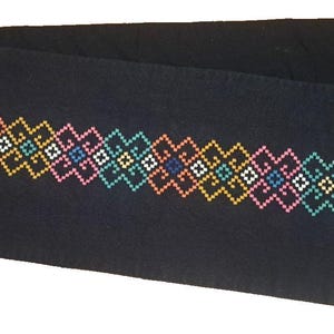 May include: A rectangular black scarf featuring a horizontal band of geometric cross-stitch embroidery. The pattern consists of repeating diamond shapes in vibrant shades of pink, yellow, white, light blue, and dark blue, with frayed edges.