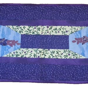 May include: A rectangular lavender-themed table runner. The runner features a central design with lavender sprigs and leaves on a light blue background. The border is a darker purple with a small floral pattern. The runner is approximately 12 inches wide and 36 inches long.
