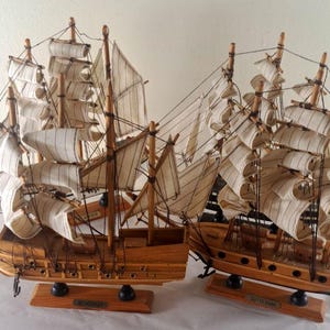 May include: Two wooden model sailing ships with detailed rigging and fabric sails. The ships are displayed on wooden bases. The sails are a cream color, and the hulls are a light brown. The ships are a classic nautical theme.