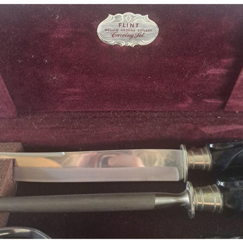 May include: A vintage Flint Hollow Ground Cutlery carving set in a burgundy velvet case. The set includes a carving knife and a fork with black handles and gold accents.