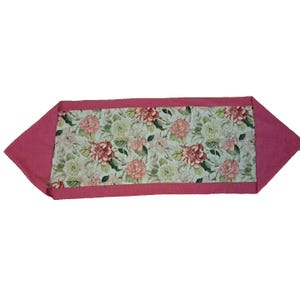 Puede incluir: Un camino de mesa floral con un borde rosa y un panel central con un estampado de flores rosas y rojas con hojas verdes sobre un fondo claro. El camino de mesa tiene forma rectangular con extremos angulados.