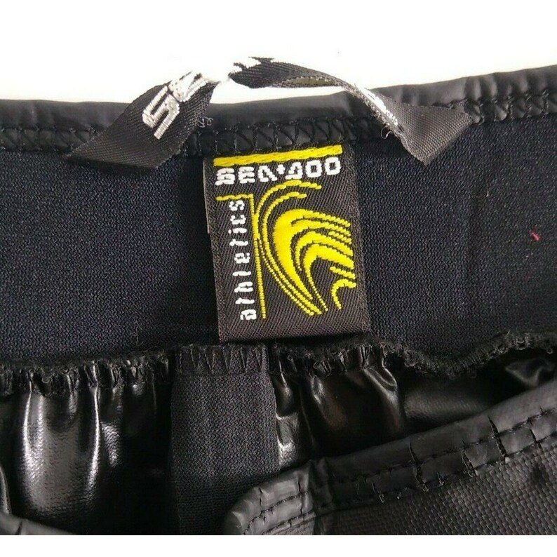 May include: Close-up of black athletic shorts with a tag that reads "SEA-DOO athletics" in white and yellow lettering. The tag features a yellow wave design. The shorts have a shiny, waterproof material.