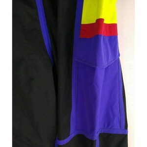 May include: Close-up of a section of black and purple athletic pants. The pants feature a color block design with yellow and red accents. A purple pocket is visible on the right side, with blue trim.