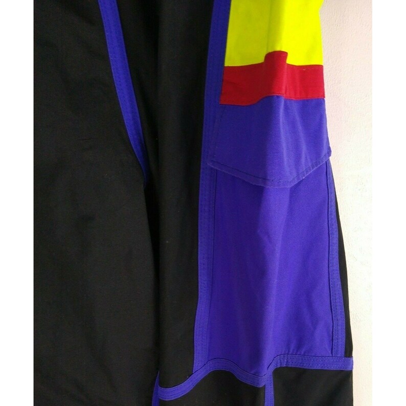 May include: Close-up of a section of black and purple athletic pants. The pants feature a color block design with yellow and red accents. A purple pocket is visible on the right side, with blue trim.