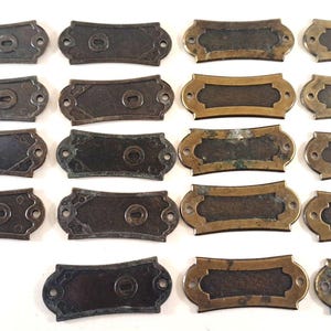 May include: A collection of antique metal nameplates in various shapes and finishes. Some are dark bronze with decorative edges, while others have a gold-toned frame. Each plate has pre-drilled holes for mounting.