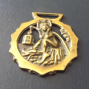 May include: A circular brass horse brass featuring a relief design of a seated imp or goblin figure holding a lantern. The figure is positioned on a textured ground with a tree stump, surrounded by a scalloped edge and a top attachment loop.