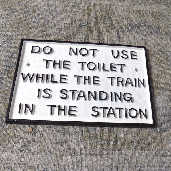 May include: A rectangular white sign with black text that reads "DO NOT USE THE TOILET WHILE THE TRAIN IS STANDING IN THE STATION." The sign has a black border and is mounted on a textured surface.