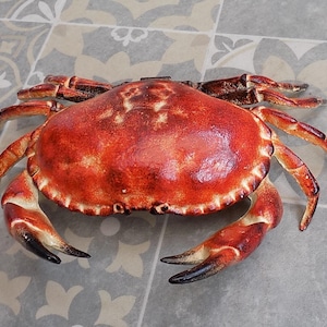 May include: A detailed, reddish-brown crab sculpture with a textured shell and multiple legs. The crab's claws have black tips. The sculpture is placed on a patterned, light-colored surface.