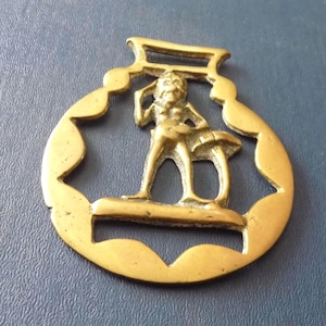 Könnte beinhalten: Ein goldfarbener Messing-Pferdeschmuck mit einem ausgeschnittenen Motiv einer stehenden Figur, die eine Hand zum Kopf führt, neben einem pilzförmigen Objekt, eingefasst in einen gewellten, kreisförmigen Rahmen mit zwei horizontalen Schlitzen.