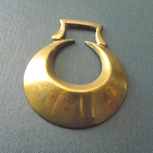 May include: A polished brass horse brass featuring a crescent moon shape with an open centre and a rectangular attachment loop at the top. The metal surface has a smooth, reflective finish with subtle variations in the golden-toned coloration.
