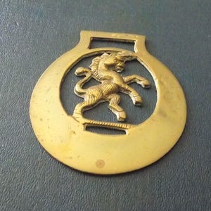 Pode incluir: Um ornamento circular de latão para cavalos com um design recortado de um cavalo empinado no centro. O objeto de metal tem um acabamento liso e polido com um laço de fixação retangular no topo e uma pequena ranhura horizontal na base.