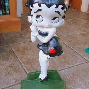 May include: A vintage Betty Boop figurine, painted in black, white, and red. The figure is posed on a green base, with a black skirt and a red heart detail. The figurine has a classic cartoon style.