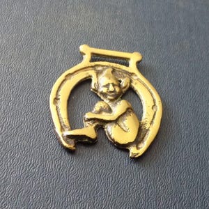 May include: A vintage brass charm featuring a seated pixie figure within a horseshoe. The charm has a loop at the top for hanging. The pixie is detailed with a pointed hat and is seated with its legs crossed. The horseshoe has nail holes.