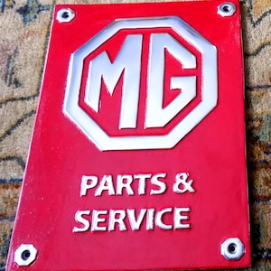 May include: A red rectangular sign featuring the silver letters "MG" within an octagonal frame. Below the logo, the words "PARTS & SERVICE" are displayed in silver. The sign is secured with four silver bolts.