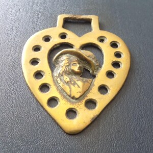May include: A heart-shaped brass horse brass featuring a central relief profile of a person wearing a wide-brimmed hat. The outer edge of the heart is decorated with a series of circular cutouts, and a rectangular loop is at the top for attachment.