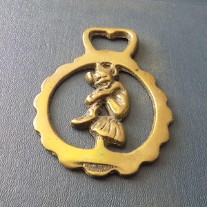 Peut inclure: Un ancien ornement de harnais en laiton représentant un lutin assis sur un champignon, encadré dans un cercle festonné. Le laiton a une boucle en haut pour l'accrocher. Le lutin est assis, les bras autour des genoux.