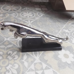 Può includere: Un ornamento per cofano Jaguar cromato a forma di giaguaro che salta. La scultura è montata su una base rettangolare nera. Un classico accessorio per auto.