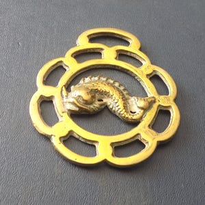 Puede incluir: Un herraje de caballo decorativo de color latón con un diseño de pez. El pez está centrado dentro de un marco circular, rodeado por un borde floral. Tiene un lazo en la parte superior para colgar.