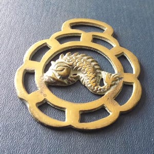 Puede incluir: Un antiguo herraje de latón para caballo con un diseño de pez detallado dentro de un marco calado. El latón tiene una pátina envejecida y dorada. El pez está finamente detallado. El marco tiene forma floral y un lazo para colgar.