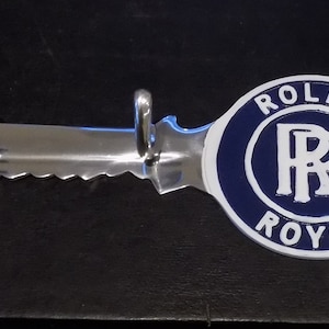 Puede incluir: Un llavero de metal pulido con dos ganchos y un borde dentado. Presenta un emblema circular con las palabras "ROLLS ROYCE" en blanco sobre fondo azul, una marca de automóviles clásica.