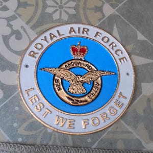May include: A circular Royal Air Force emblem with a blue center, featuring a gold eagle and a red crown. The words "Royal Air Force" and "Lest We Forget" are inscribed around the edge in white lettering. The emblem has a white border with gold accents.