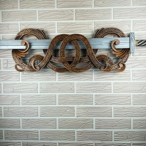 May include: A wooden sword holder with intricate Celtic knotwork design. The holder is brown and features two circular end pieces and a central knot design. A silver sword is placed through the holder, creating a decorative display against a brick wall.