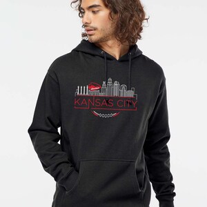 May include: A black hooded sweatshirt featuring a red and white graphic of the Kansas City skyline. The design includes the words "KANSAS CITY" in a red font, with a football-shaped element below the text. The sweatshirt has a front pocket and a drawstring hood.
