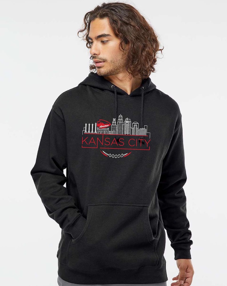 May include: A black hooded sweatshirt featuring a red and white graphic of the Kansas City skyline. The design includes the words "KANSAS CITY" in a red font, with a football-shaped element below the text. The sweatshirt has a front pocket and a drawstring hood.
