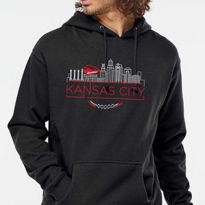 May include: A black hooded sweatshirt featuring a red and white graphic of the Kansas City skyline. The text "KANSAS CITY" is displayed in red below the skyline design. The sweatshirt has a front pocket and a drawstring hood.