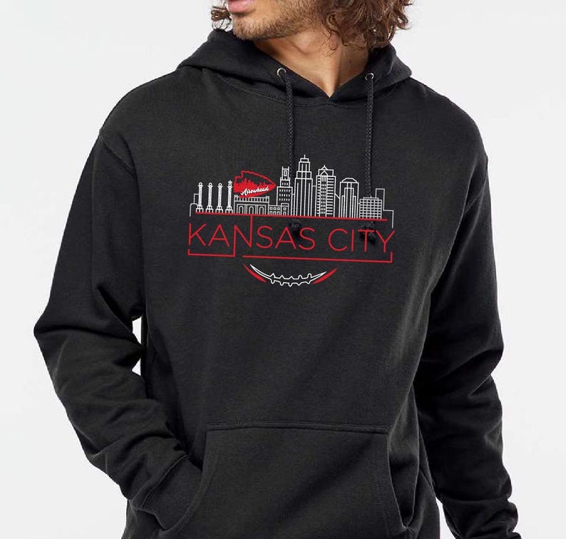 May include: A black hooded sweatshirt featuring a red and white graphic of the Kansas City skyline. The text "KANSAS CITY" is displayed in red below the skyline design. The sweatshirt has a front pocket and a drawstring hood.