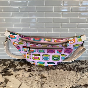 May include: A pink fanny pack with a white strap and a pattern of colourful canned goods with fish bones on the front. The fanny pack has three zipped compartments.