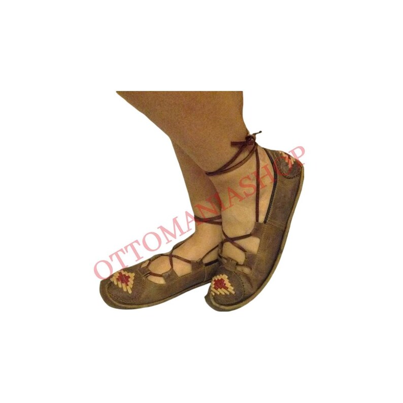 May include: Brown leather shoes with laces and red and white embroidery. The shoes are tied around the ankles.