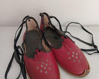 May include: A pair of red leather sandals with black straps and a decorative cut-out design. The sandals have a rounded toe and a dark brown sole. The laces are long and black.