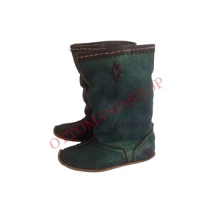K&ouml;nnte beinhalten: Ein Paar gr&uuml;ne Lederstiefel mit einem dekorativen Stichdetail am oberen Rand und einem kleinen Kreuzmuster auf der Vorderseite. Die Stiefel haben eine flache Sohle und sind aus weichem, geschmeidigem Leder gefertigt.