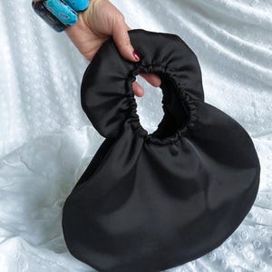 Op de afbeelding: Een zwarte satijnen handtas met een ronde handgreep. De tas heeft een geplooid ontwerp en een afgeronde vorm. De handgreep is een grote, open cirkel. De tas wordt tegen een witte achtergrond gehouden.